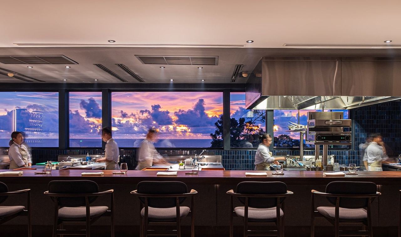 The chef’s counter at PRU, with the open kitchen and a sunset sky over the sea through the windows