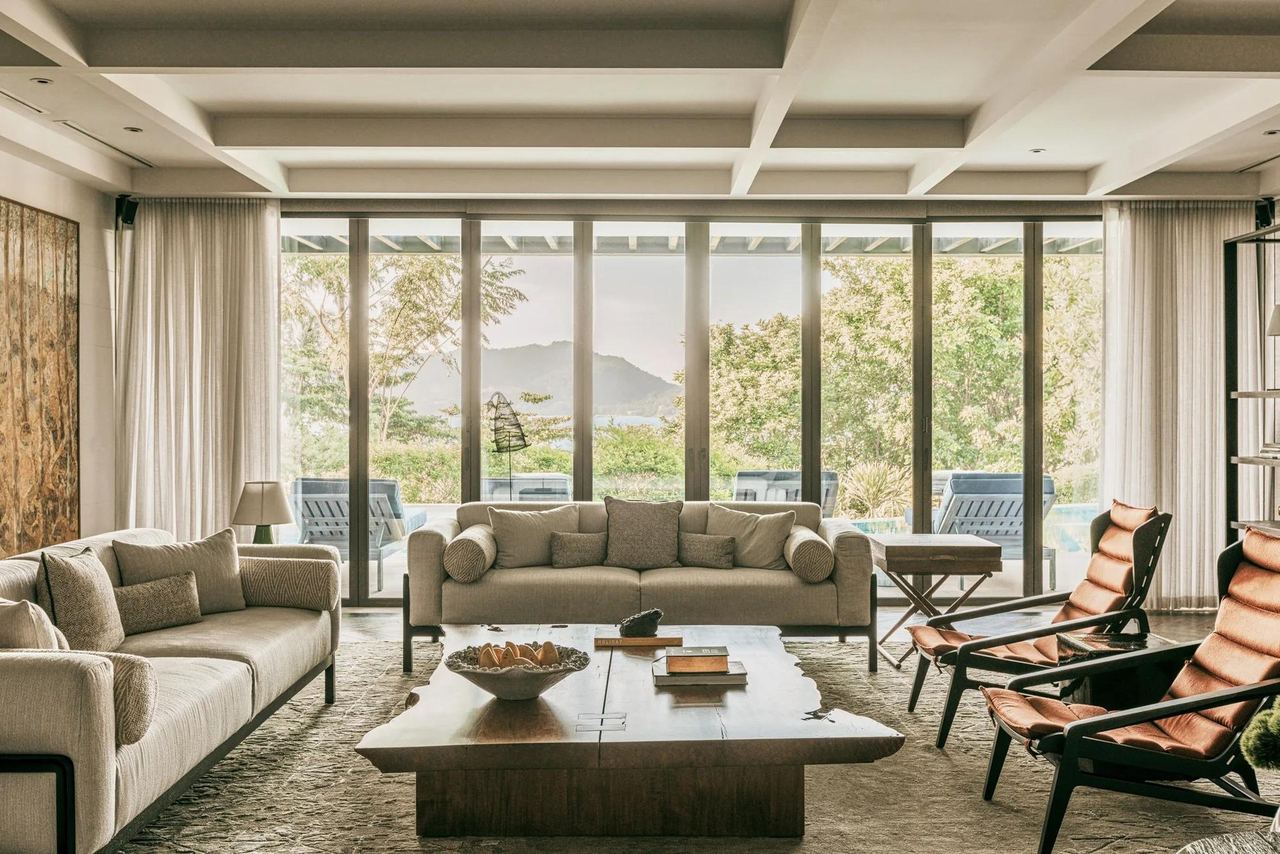 The sitting room of The Palm House at Rosewood Phuket, with a hill view through tall windows